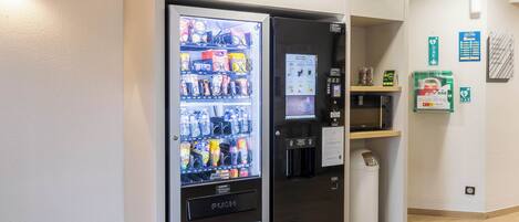 Máquina expendedora con aperitivos y bebidas, un microondas y un cubo de basura en un espacio limpio y moderno.