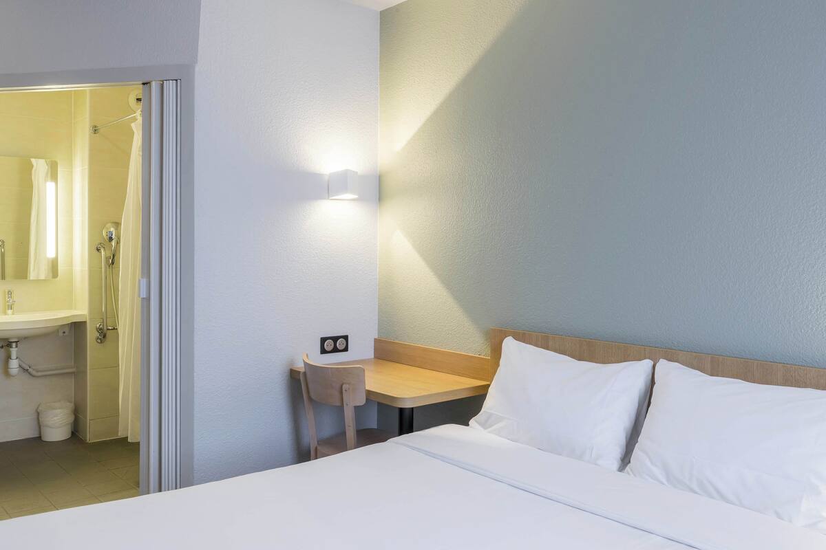A hotel room with a bed, a bedside table, a chair, a bathroom with a sink, and a mirror.