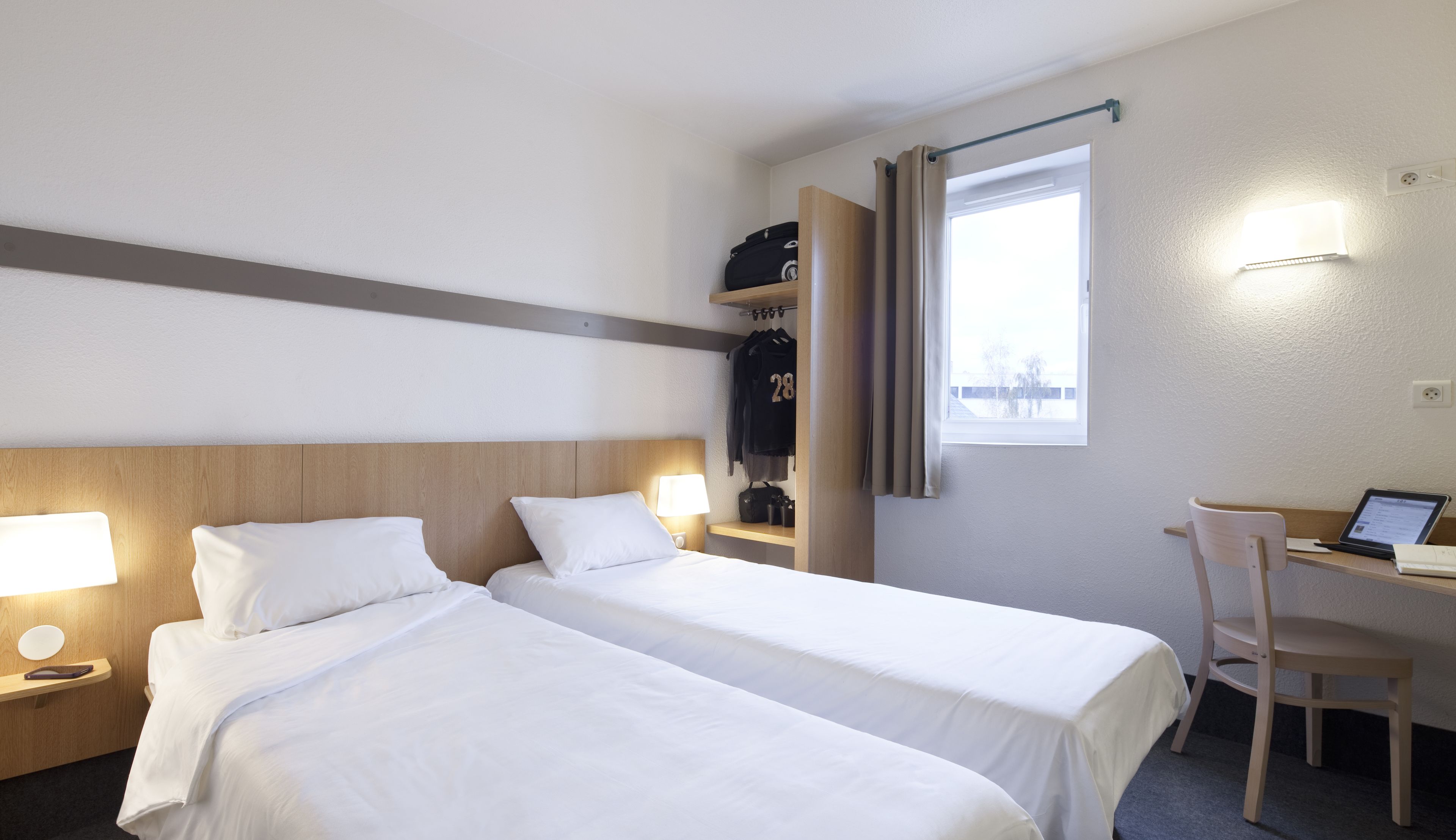 A hotel room with two beds, a desk with a laptop, a chair, and a window with curtains.