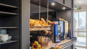 Una barra de desayuno con pan, bollería y fruta. Hay cestas, frascos y una máquina de café.