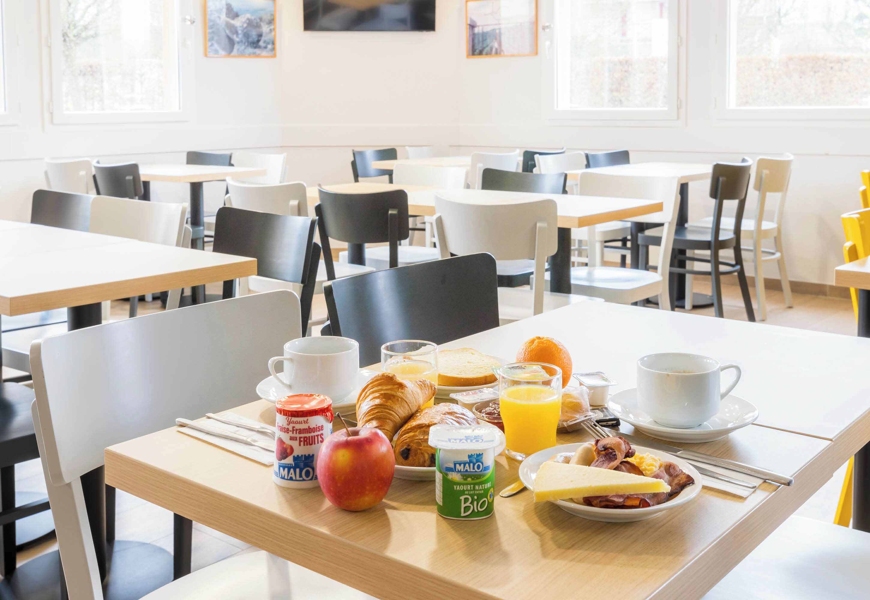 Una mesa de desayuno con croissants, frutas, yogur y jugo en un comedor luminoso con varias mesas y sillas.