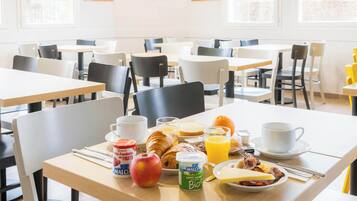 Una mesa de desayuno con croissants, frutas, yogur y jugo en un comedor luminoso con varias mesas y sillas.