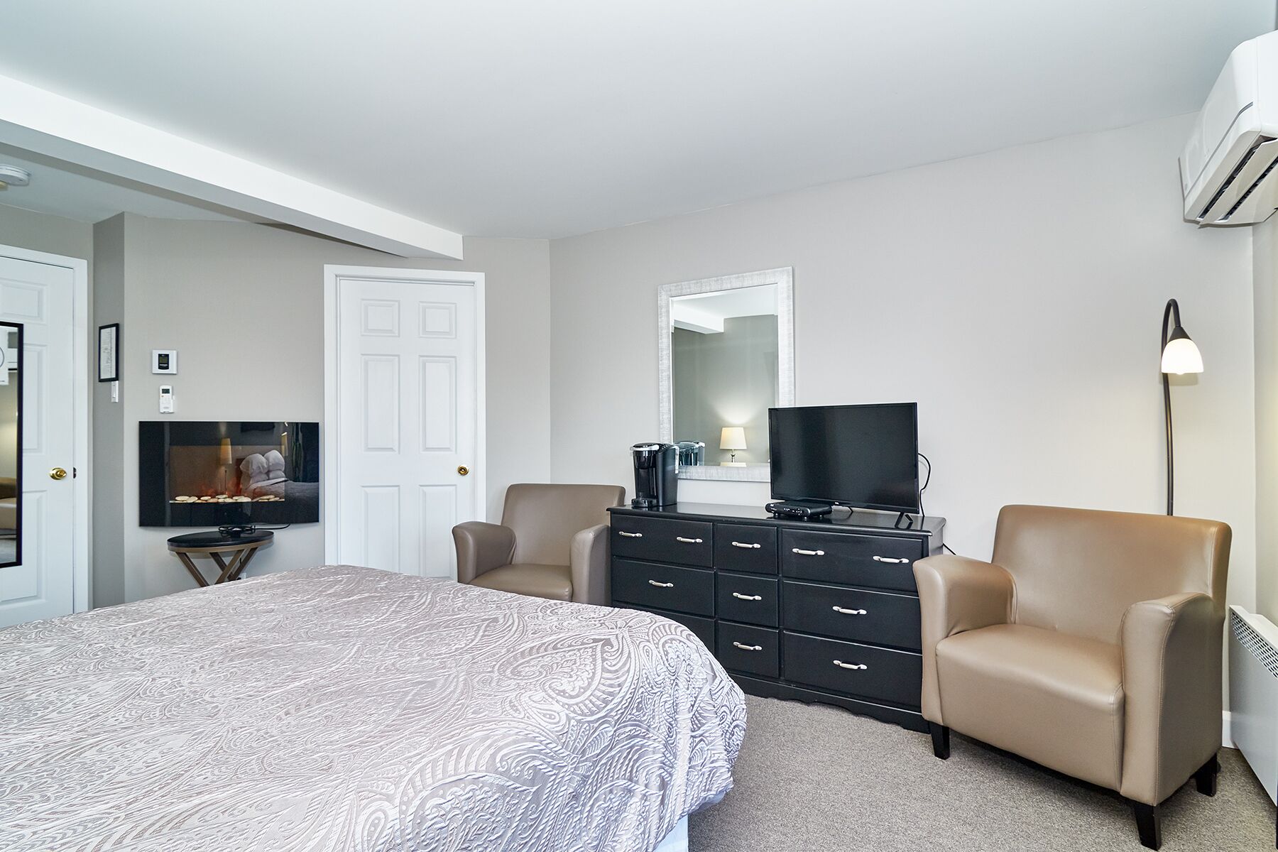A bedroom with a bed, two armchairs, a dresser, a television, a mirror, and an air conditioning unit.