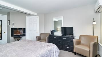 A bedroom with a bed, two armchairs, a dresser, a television, a mirror, and an air conditioning unit.