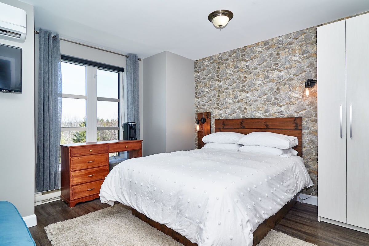 Une chambre avec un mur en pierre, un cadre de lit en bois, un bureau avec une chaise, une armoire et une fenêtre avec des rideaux.