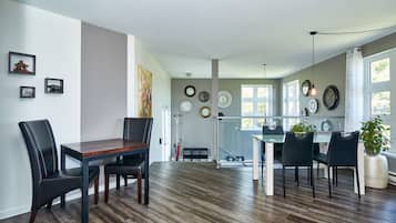 A spacious dining area with a wooden table and chairs, a kitchen in the background, and a large window providing natural light.