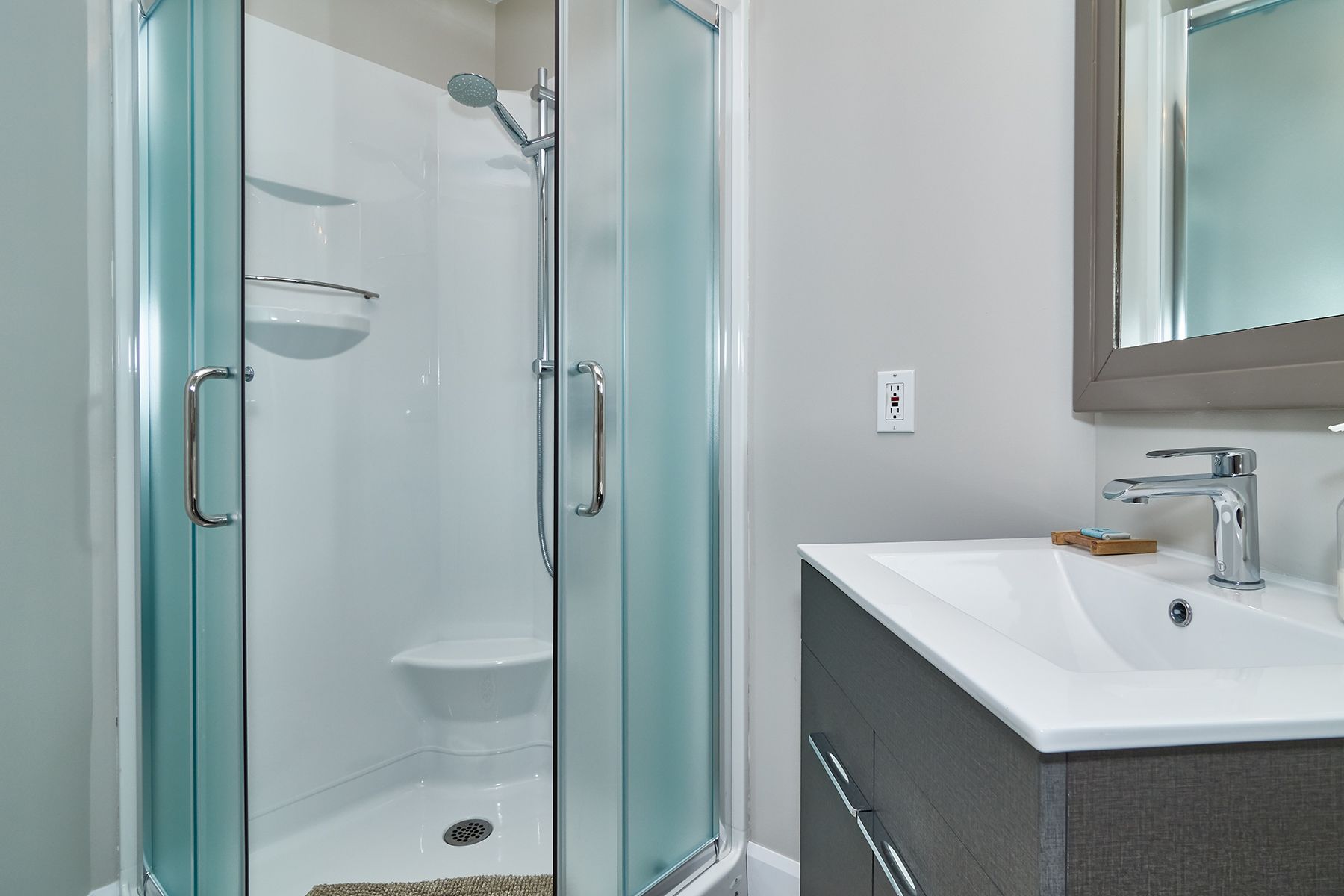 Une salle de bain moderne avec une douche à l’italienne en verre, un lavabo blanc avec un miroir et un meuble-lavabo à carreaux foncés.