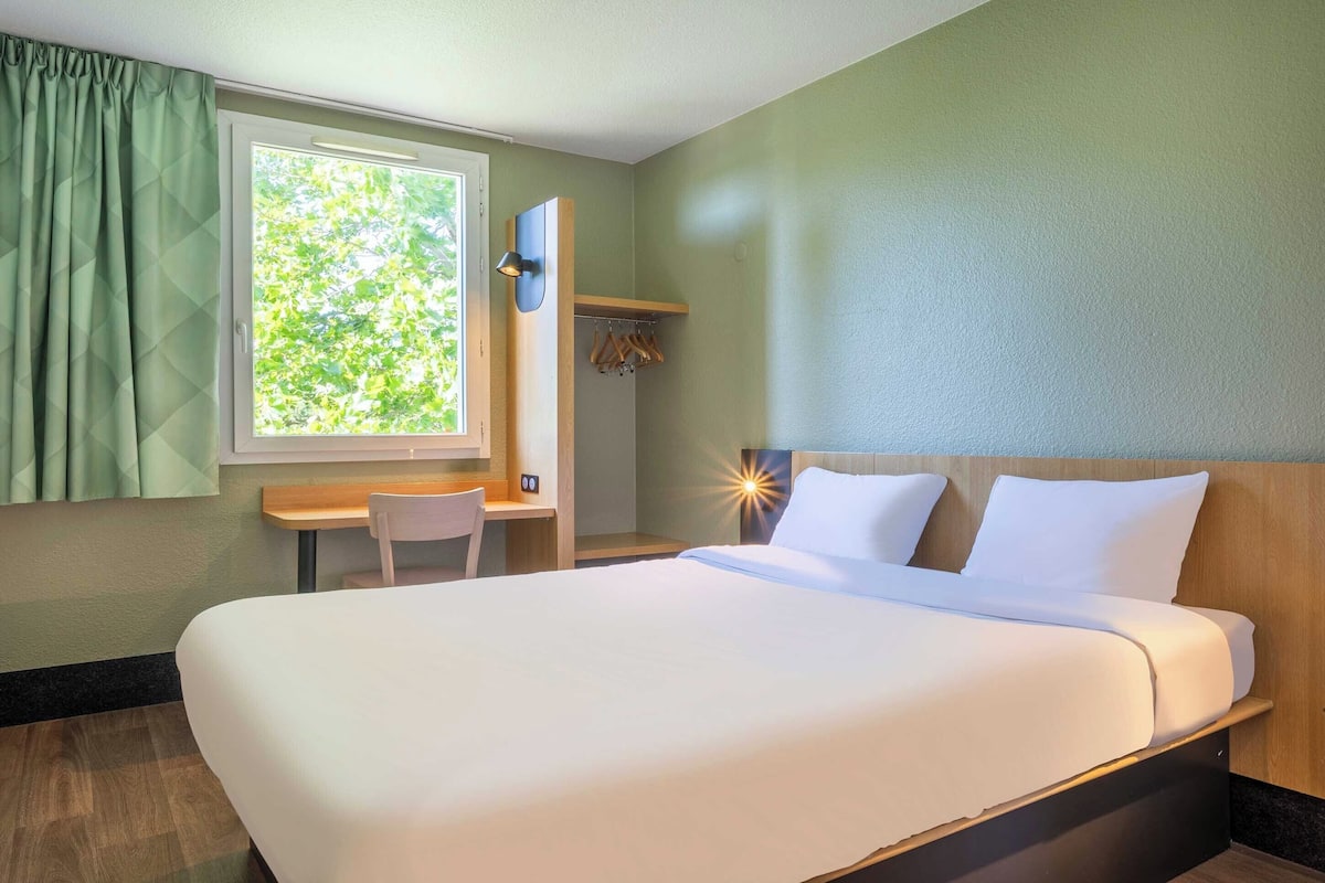Ein Hotelzimmer mit einem großen Bett, einem Schreibtisch, einem Sessel und einem fenster mit Vorhängen.
