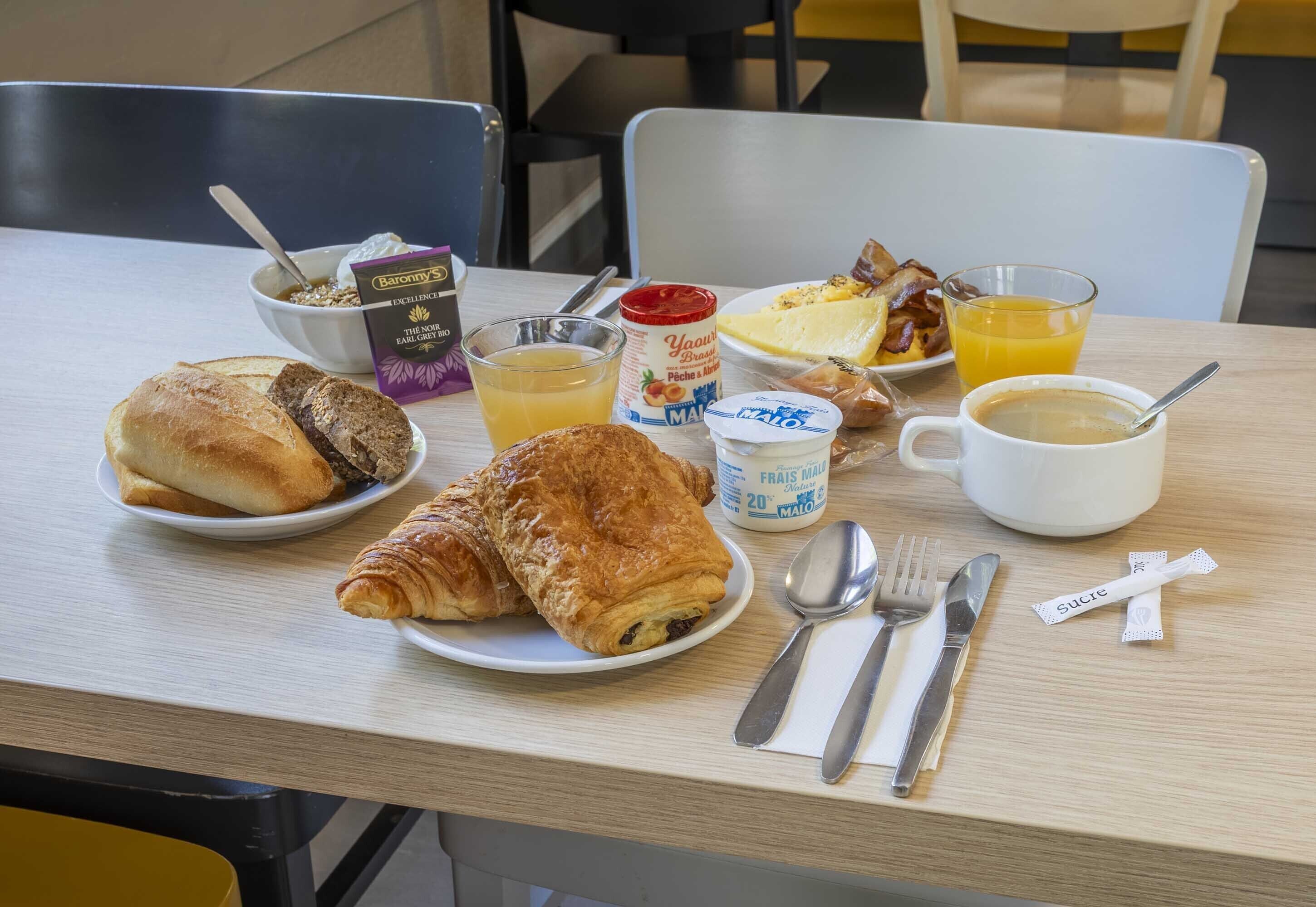 A breakfast table with croissants, a bowl of cereal, a glass of orange juice, a plate of bacon, a cup of coffee, and a packet of sugar.