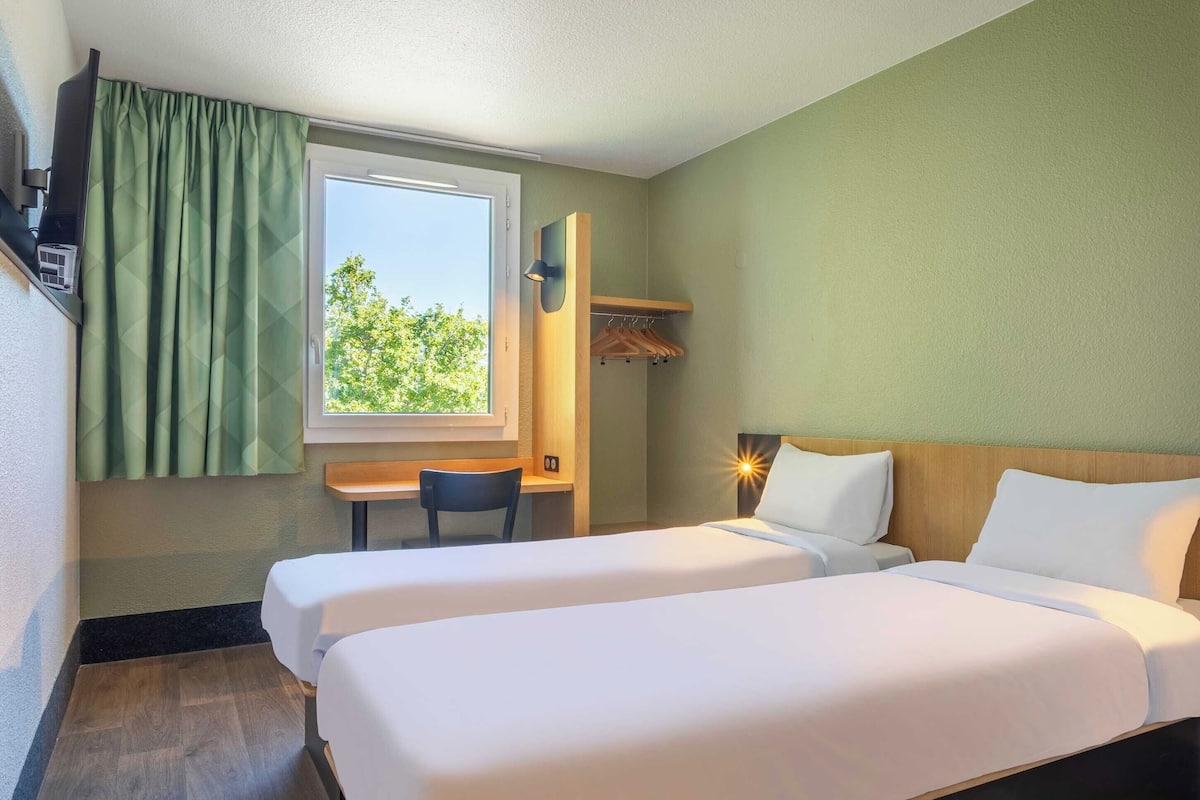 Ein Hotelzimmer mit zwei Betten, einem Schreibtisch, einem Stuhl, einem Fenster mit Vorhängen und einem an der Wand befestigten Regal.