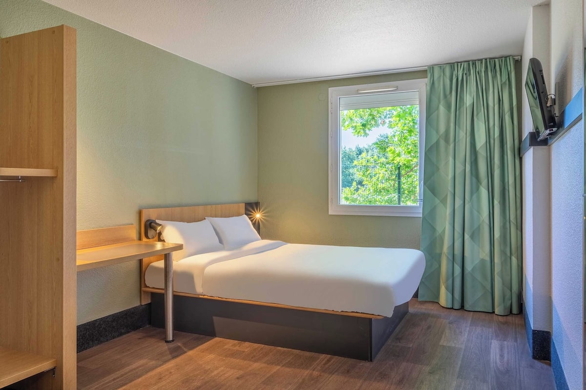 Ein Hotelzimmer mit Bett, Nachttisch, einem Fenster mit Vorhängen und einem an der Wand montierten Fernseher.