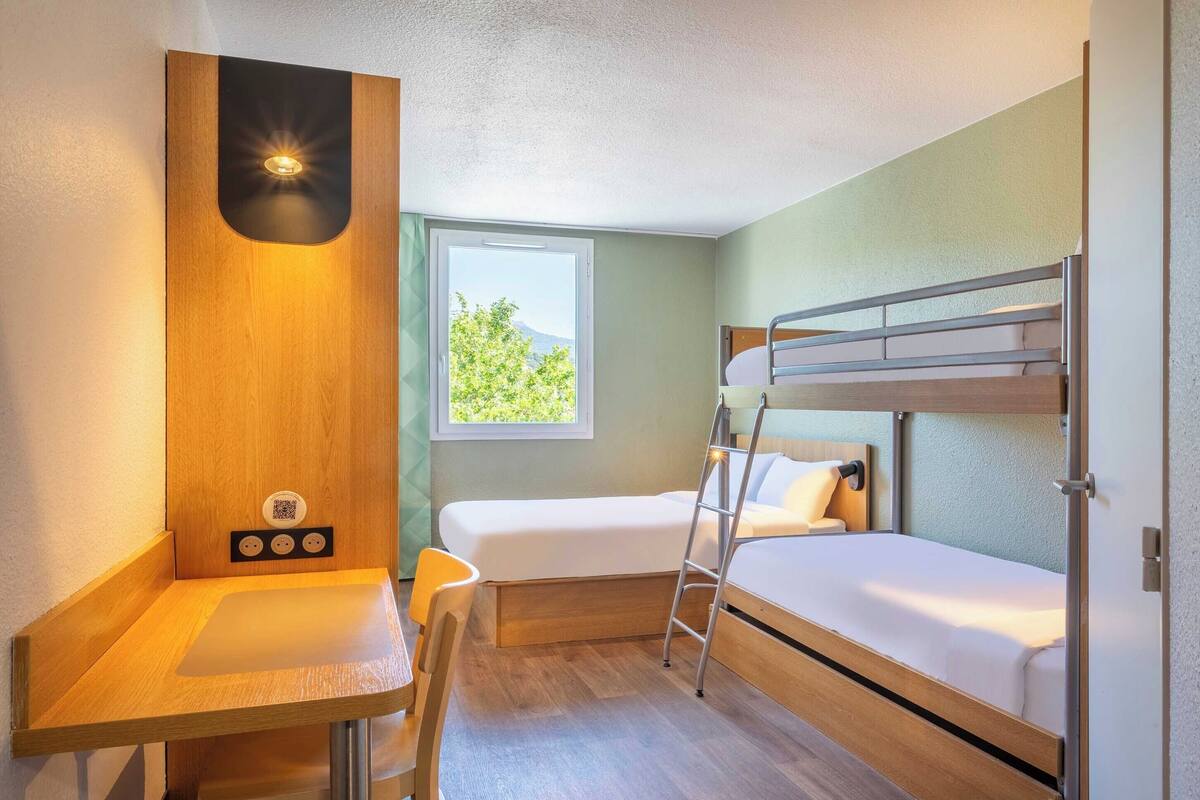 Ein kleines Zimmer mit einem Etagenbett, einem Schreibtisch mit Stuhl, einem Fenster mit Ausblick und einer Wandleuchte.