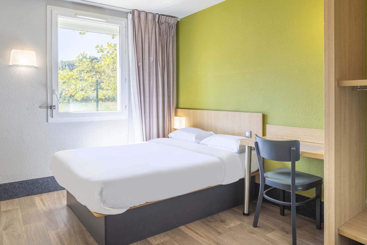 Ein Hotelzimmer mit einem Bett, einem Schreibtisch, einem Stuhl, einem Fenster mit Vorhängen und einer Lampe.