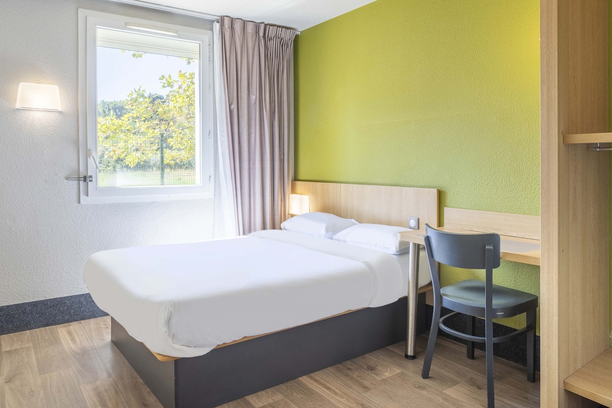Una habitación de hotel con cama, escritorio, silla, ventana con cortinas y lámpara.