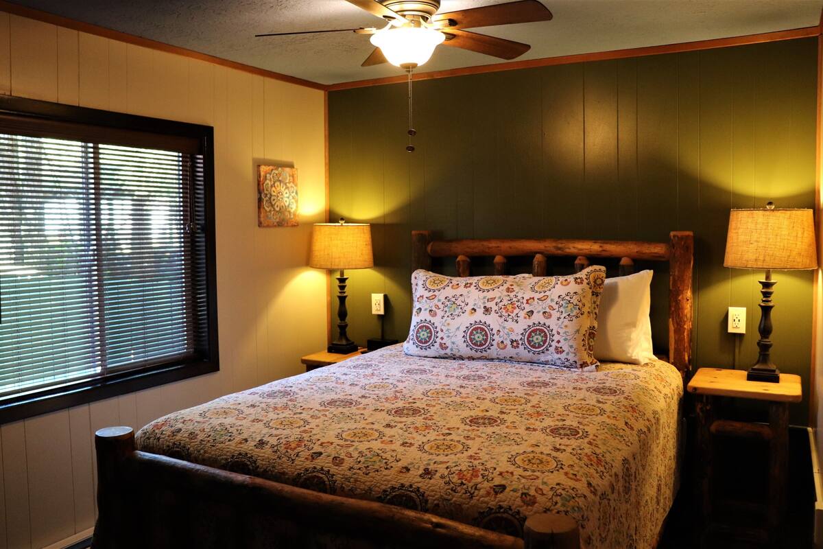 A bedroom with a bed, two bedside lamps, a ceiling fan, and a window with blinds.