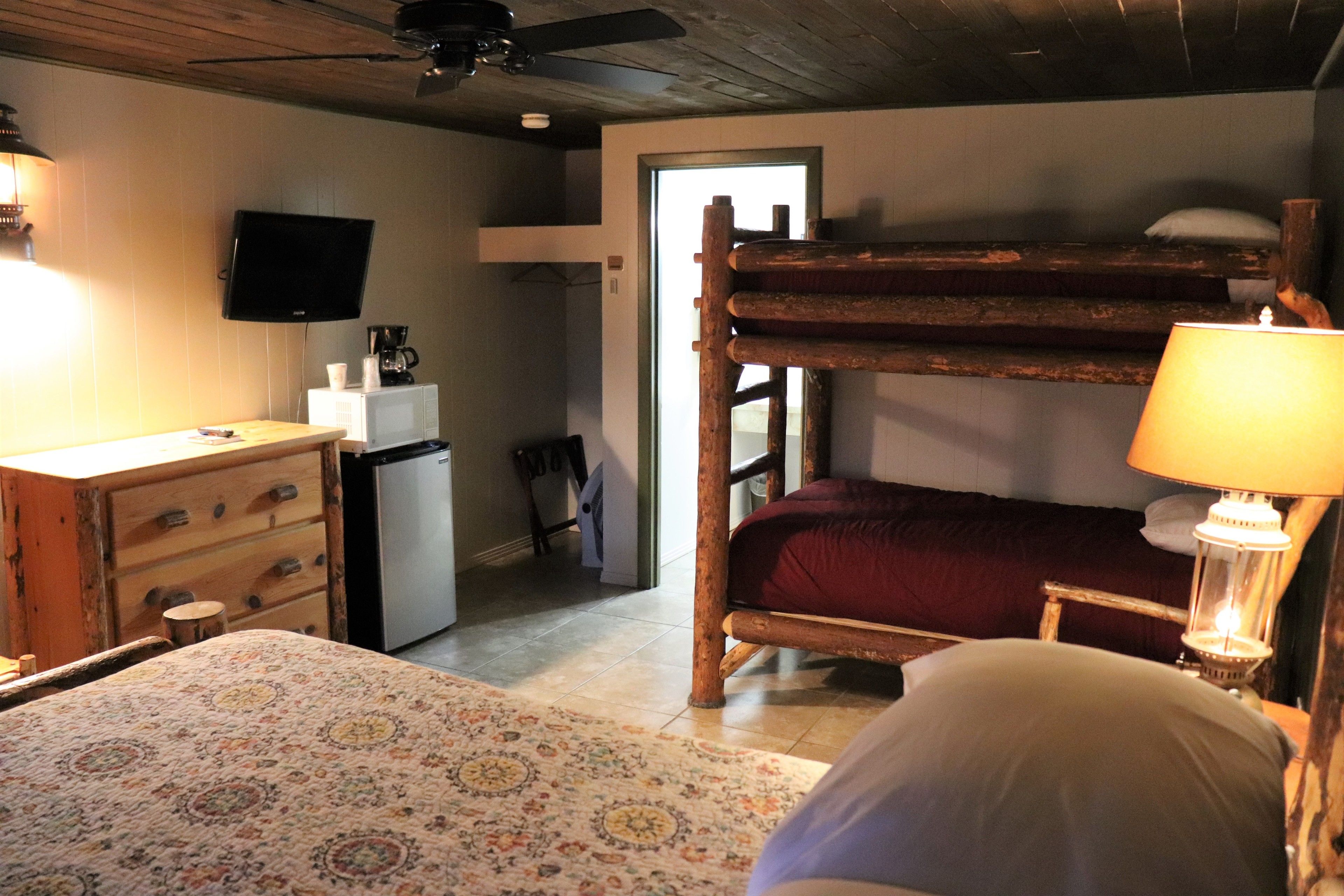 Una habitación rústica con una cama litera, un escritorio con cajones, un refrigerador pequeño, un televisor montado en la pared y una lámpara en la mesita de noche.