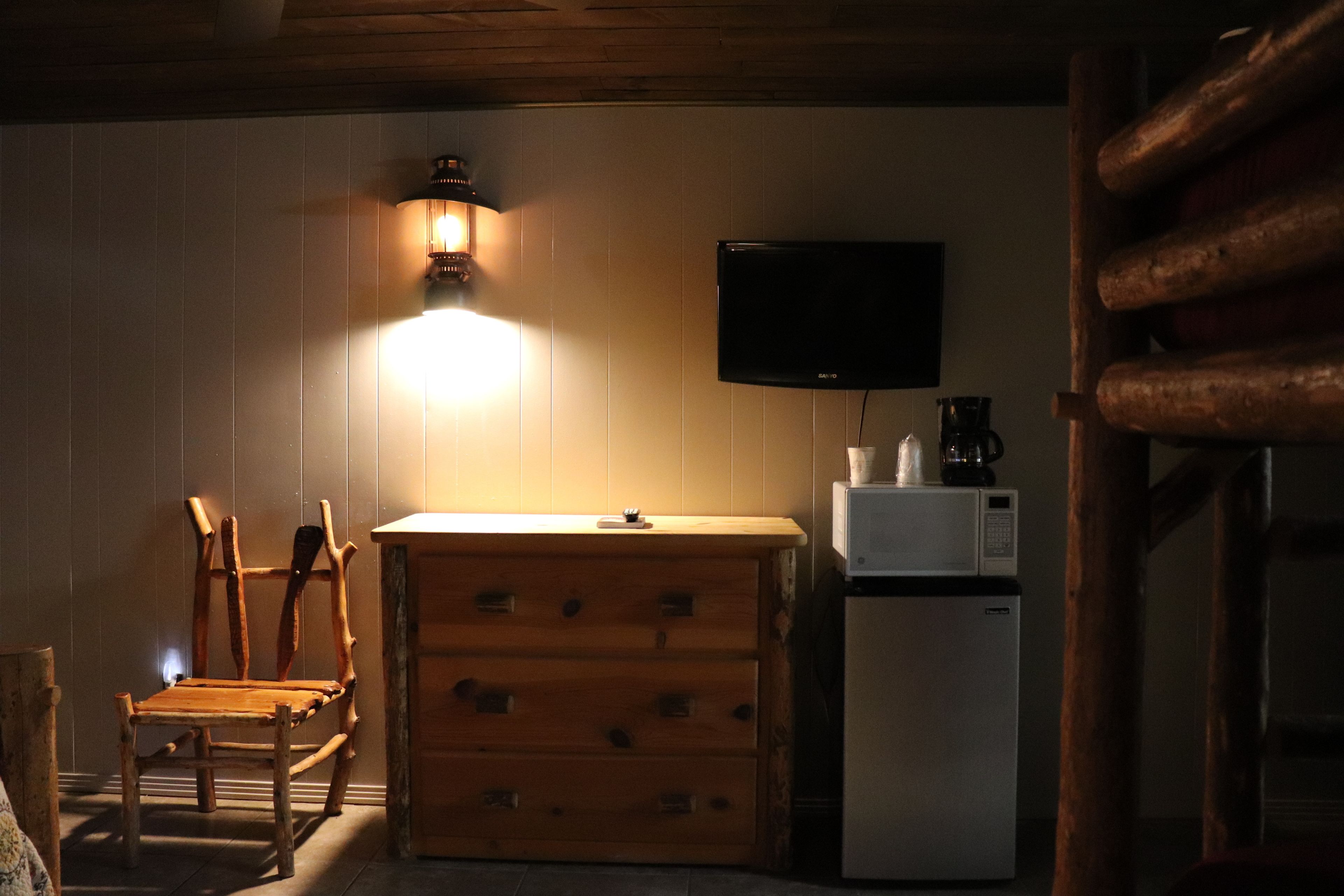 A cozy room with a wooden chair, a wooden dresser, a white refrigerator, and a flat-screen TV mounted on the wall.