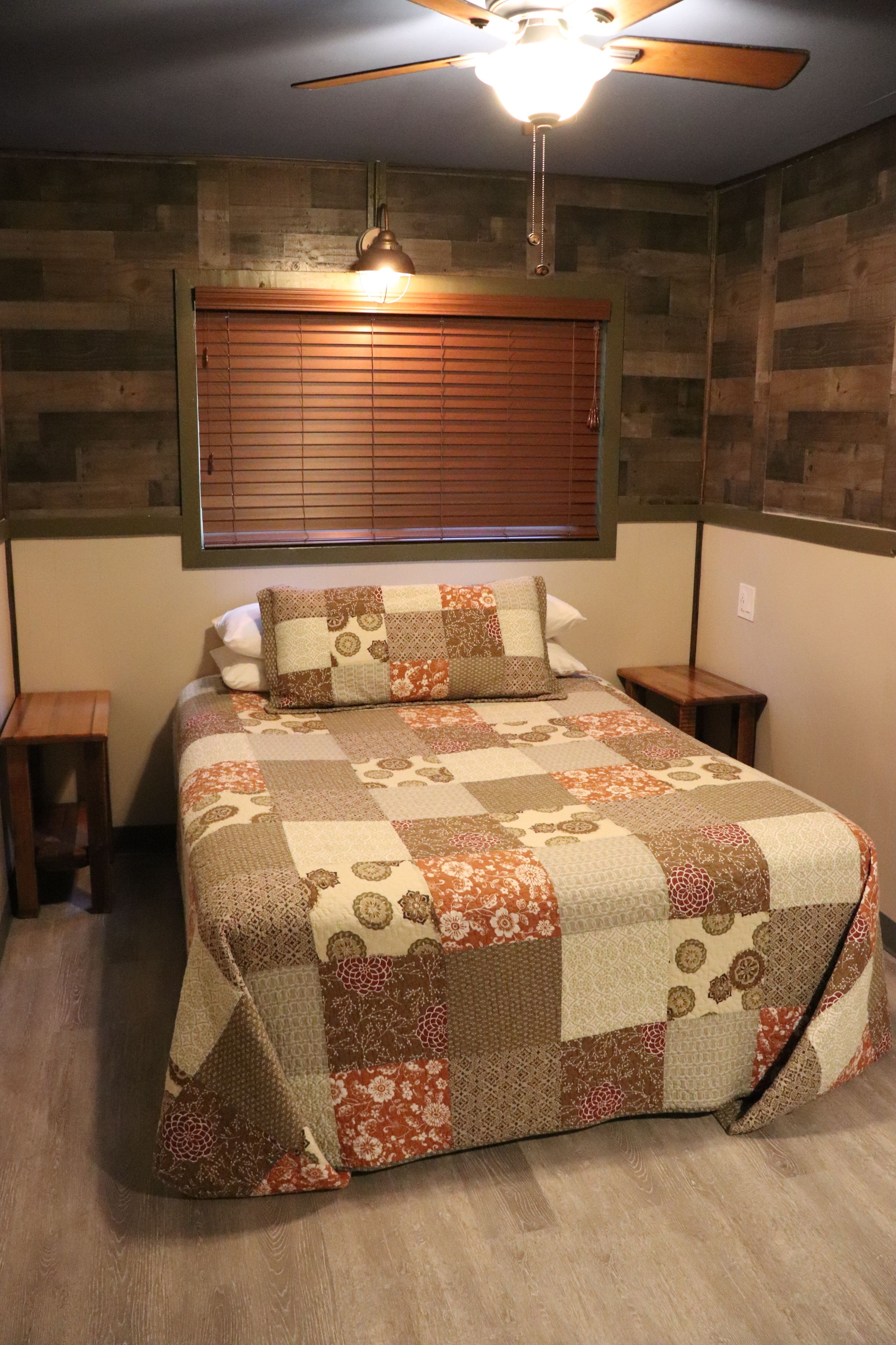 A bedroom with a quilted bedspread, a ceiling fan, a window with blinds, and a bedside table.