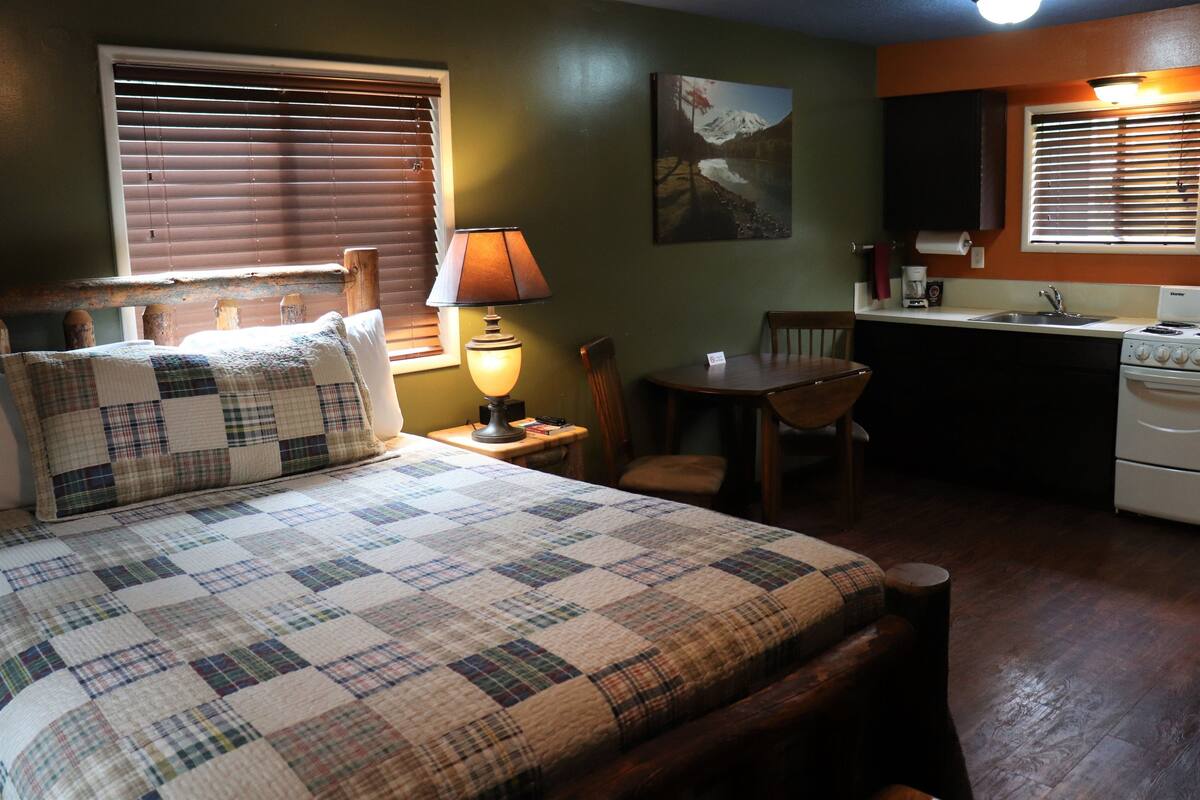 A cozy room with a bed, a desk, a chair, a lamp, a painting, and a kitchen area.