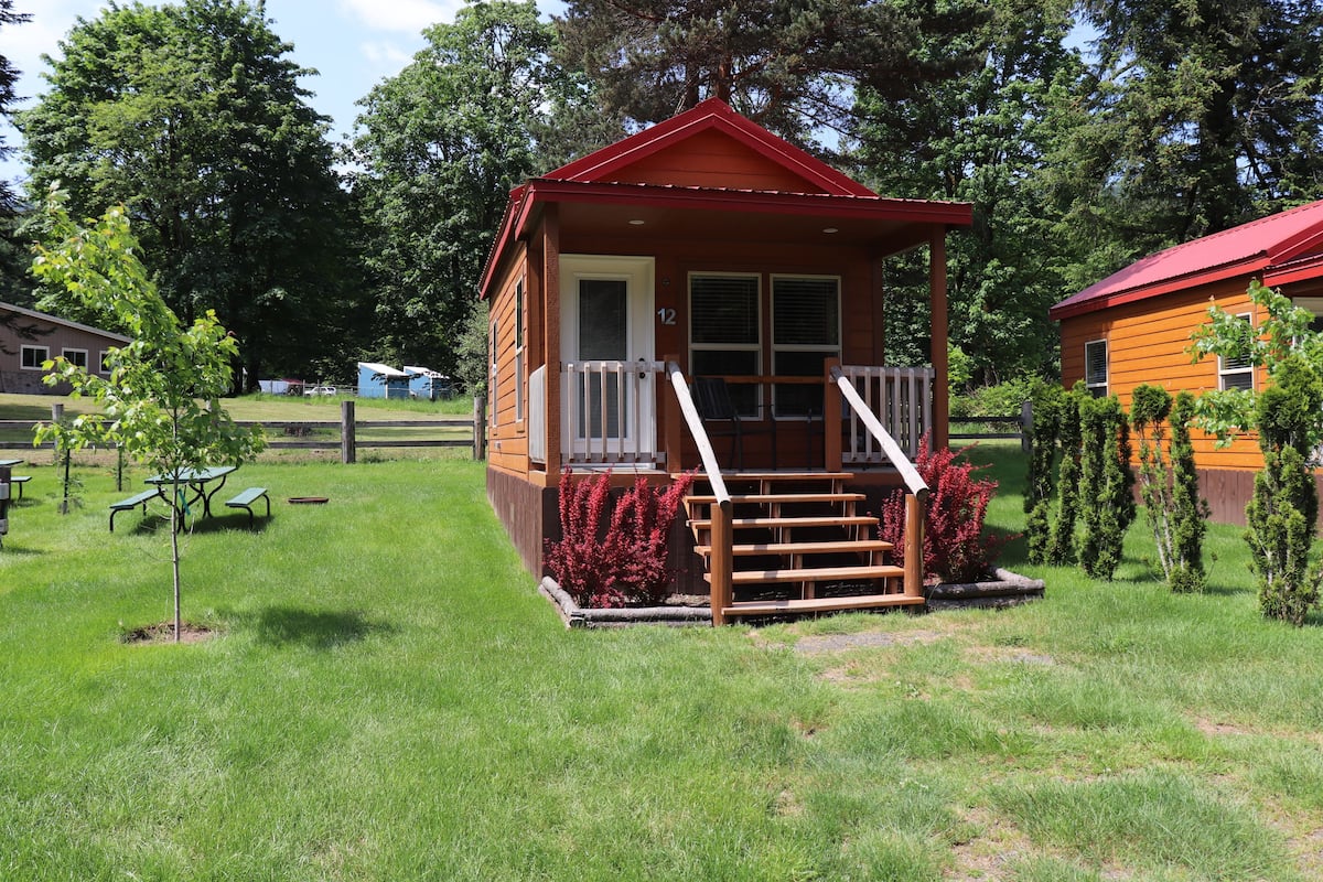 A small wooden cabin with a red roof, white porch railing, and a set of stairs leading to the entrance.