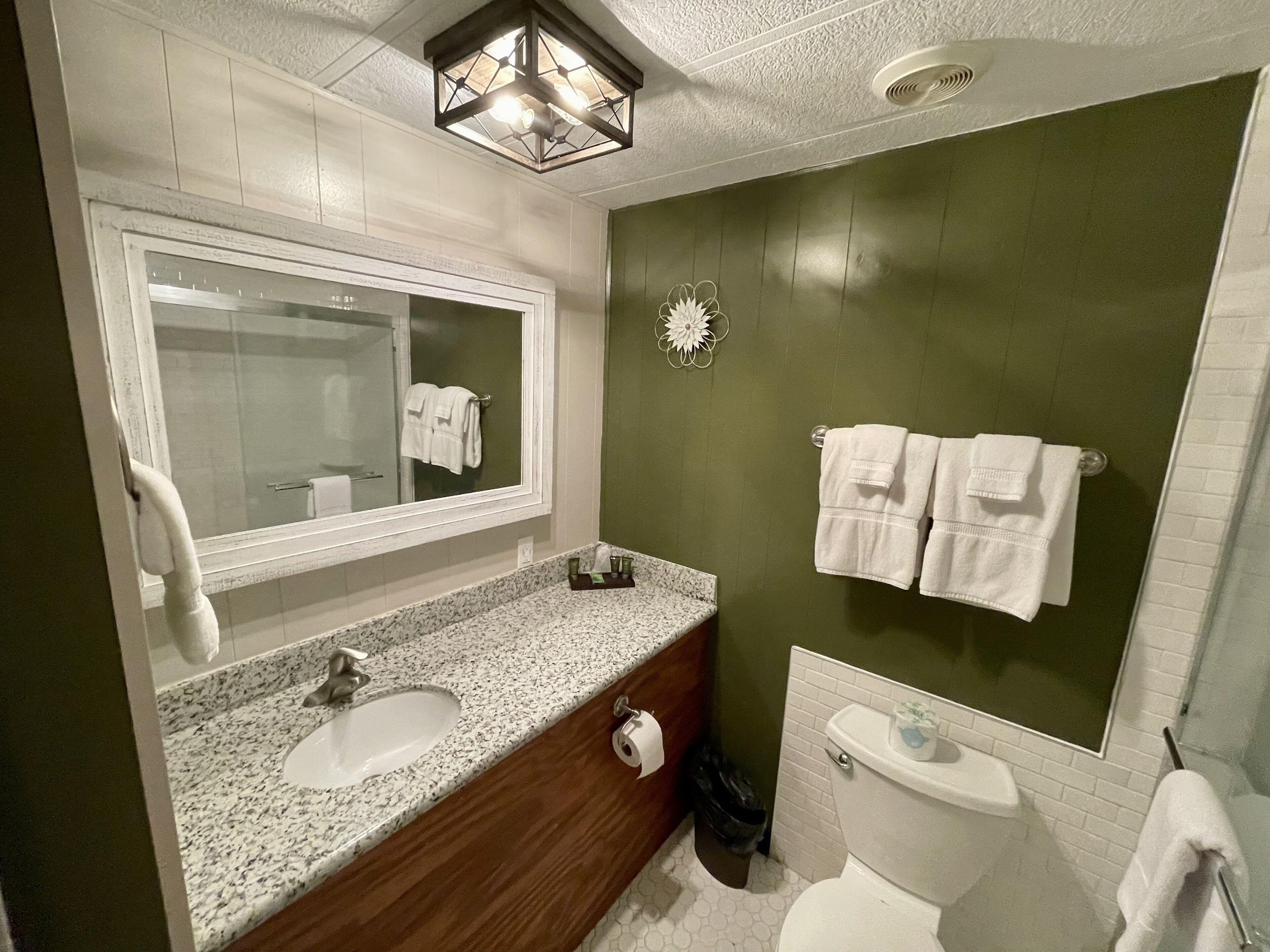 Ein Badezimmer mit Marmorwaschtisch, einem Holzwaschtischunterschrank, einem großen Spiegel, einer Toilette und einer grünen Akzentwand.