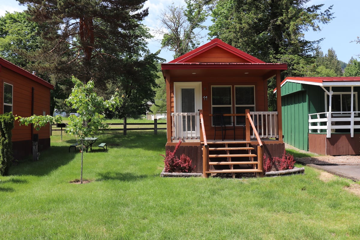 A small cabin with a red roof, a porch with wooden steps, and a white railing.