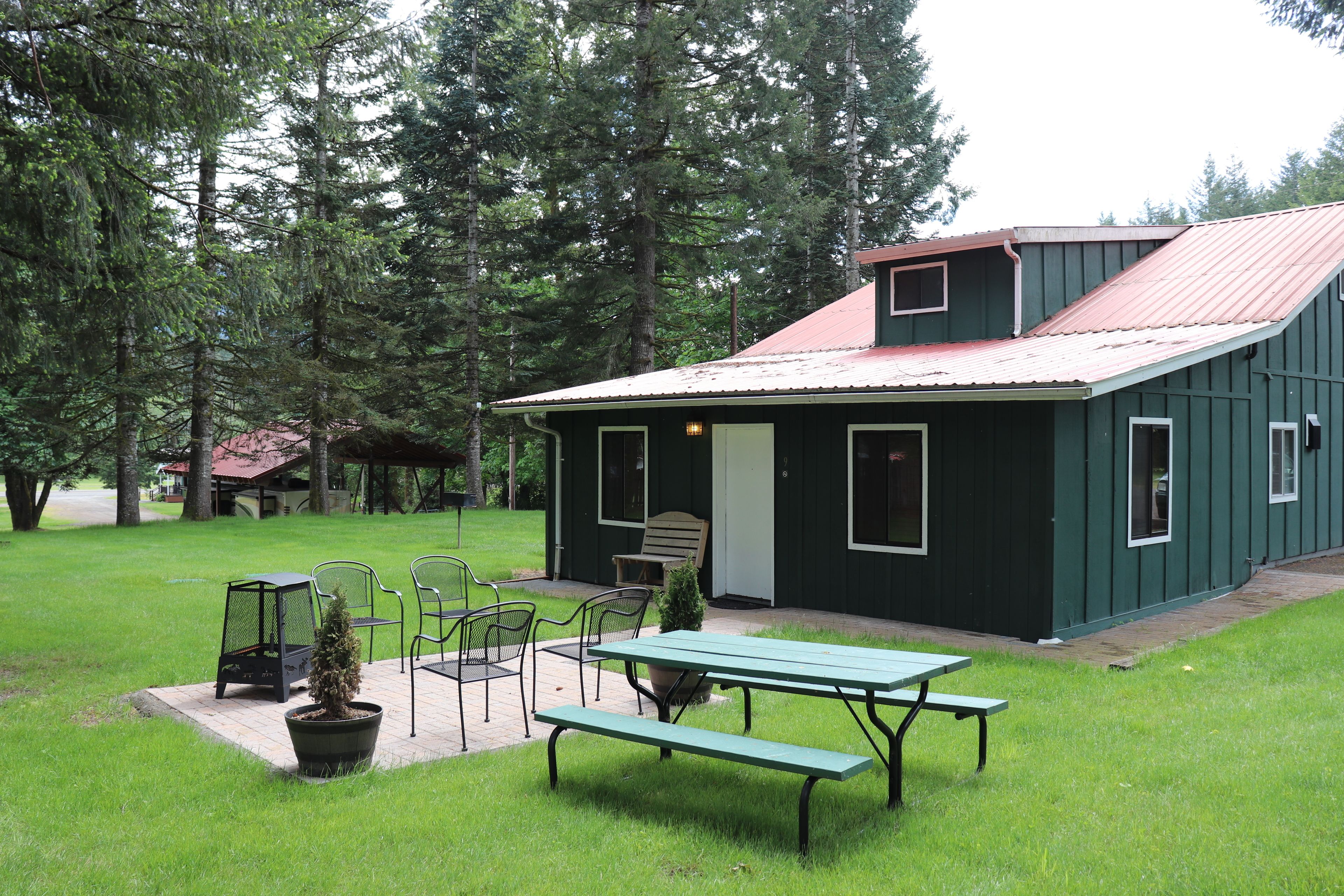 A green building with a red roof, surrounded by trees, featuring a picnic table and chairs, and a small porch area.
