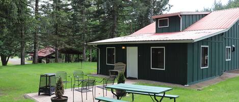 A green building with a red roof, surrounded by trees, featuring a picnic table and chairs, and a small porch area.