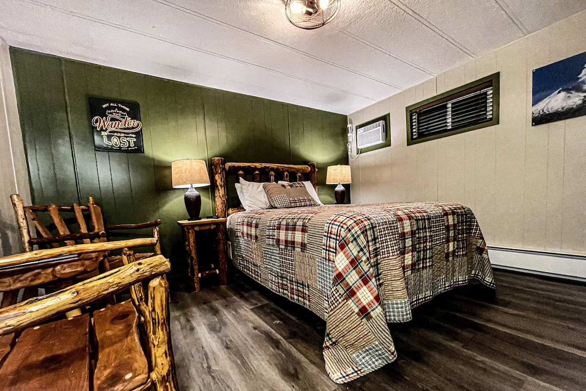 A bedroom with a wooden bed, two bedside tables with lamps, a wooden chair, a green wall, and a picture of a mountain.