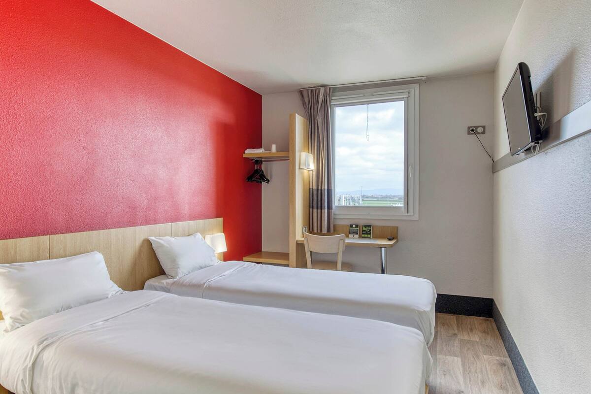 Habitación de hotel con dos camas, un escritorio, una silla, un televisor montado en la pared y una ventana con cortinas.