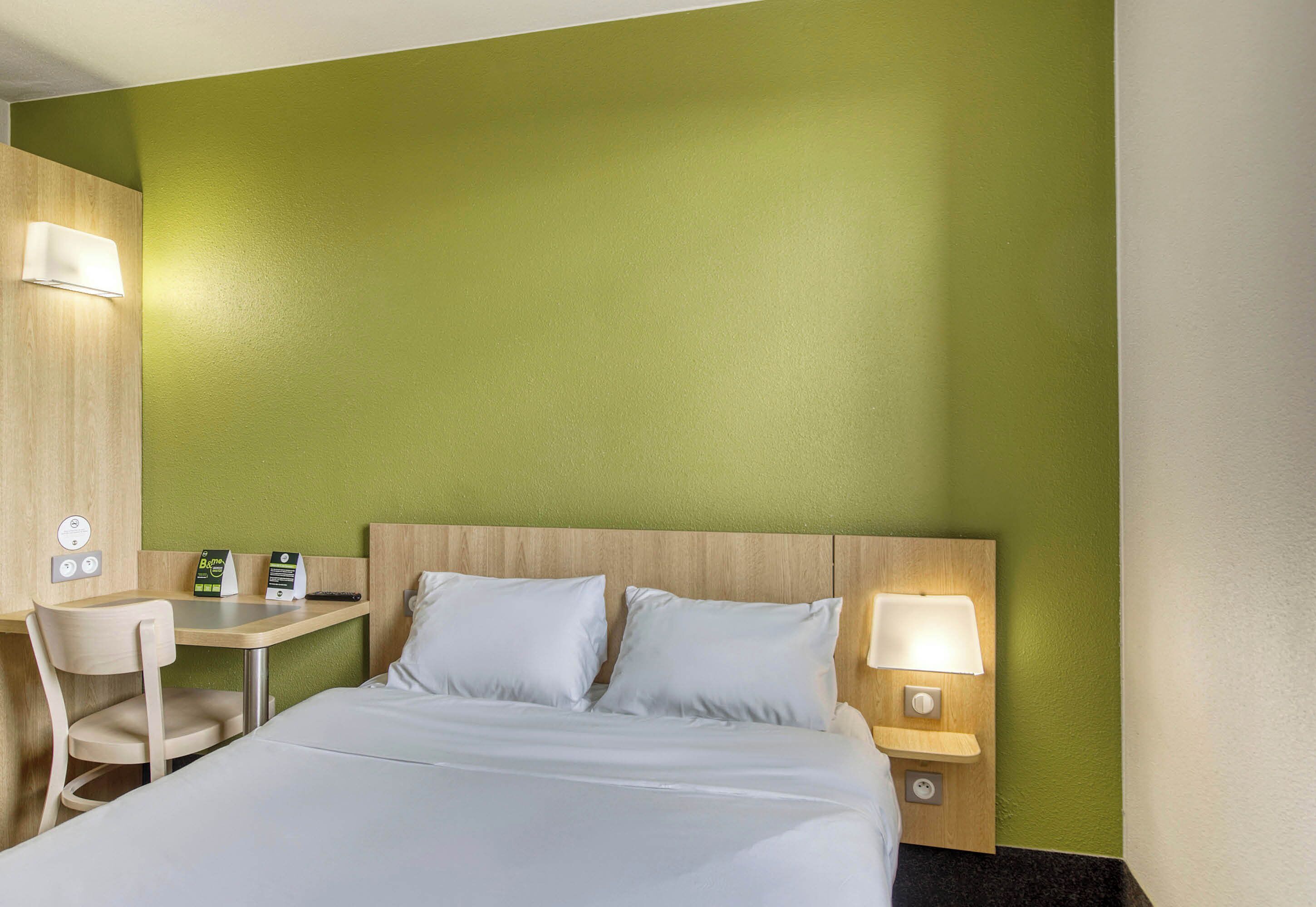 A hotel room with a bed, a desk, a chair, and a nightstand with a lamp.