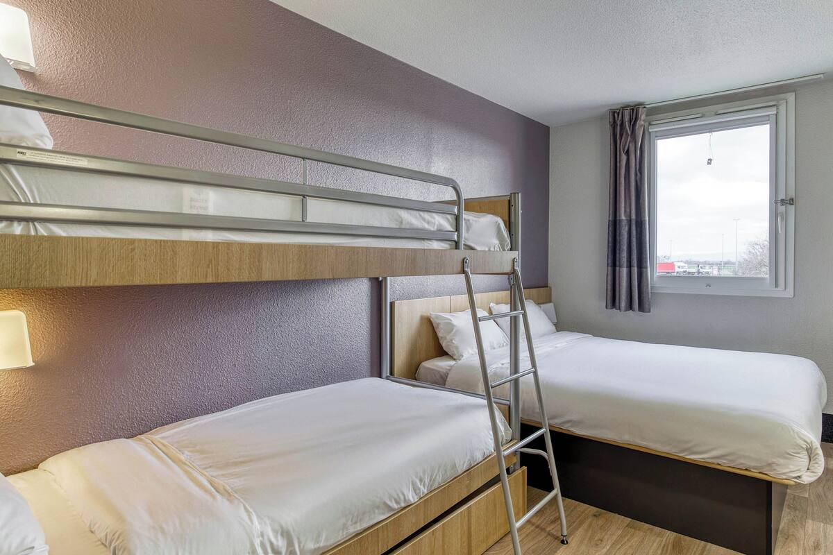 Habitación de hotel con dos camas individuales, una litera, una ventana con cortinas y una lámpara fijada a la pared.