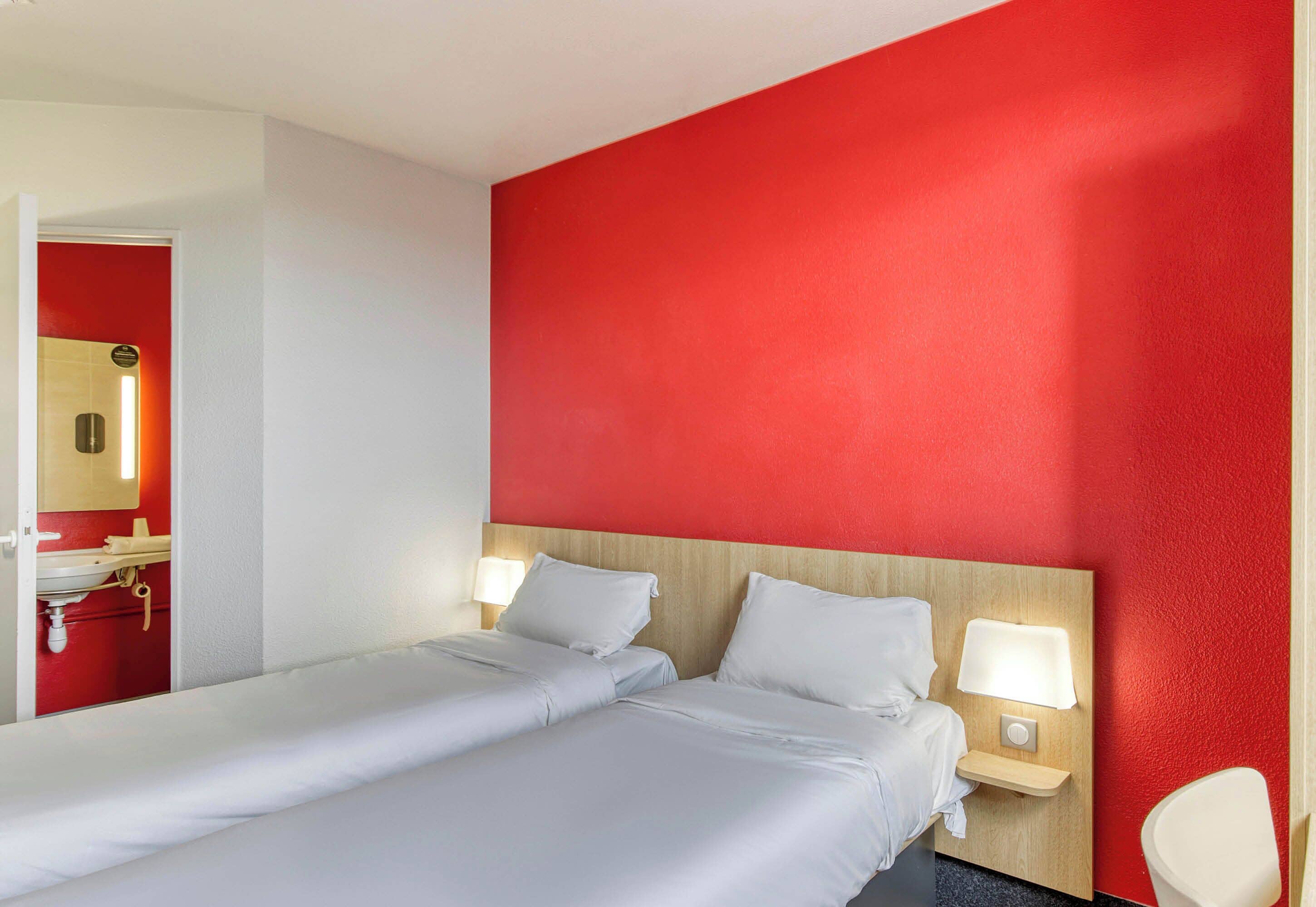 A hotel room with a large bed, two bedside tables with lamps, a red accent wall, and a bathroom visible through an open door.