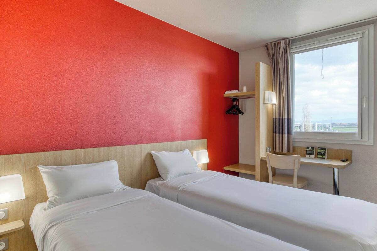 Habitación de hotel con dos camas, un escritorio, una silla, una ventana con cortinas y vistas a la ciudad.