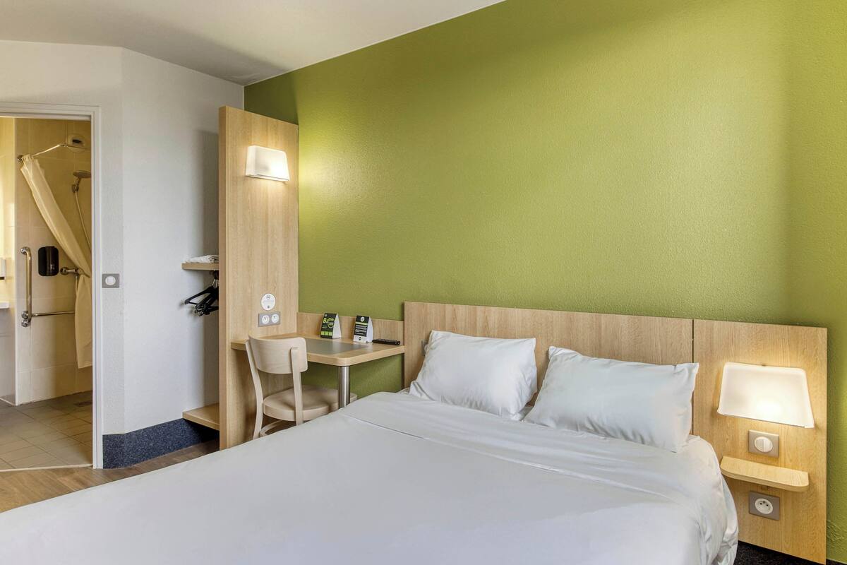 Habitación de hotel con cama, escritorio, silla, zona de ducha y pared verde.
