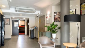 A hotel lobby with a reception desk, a seating area with a chair and a small table, a potted plant, and a large pillar. - B&B HOTEL Avignon (2) (Le Pontet)