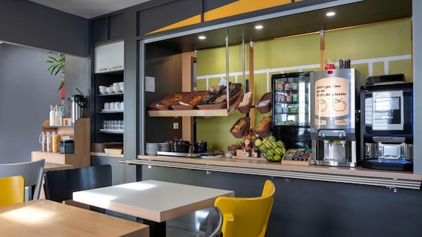 A café with a counter displaying pastries, a coffee machine, and a menu board. - B&B HOTEL Avignon (2) (Le Pontet)