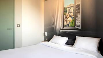 Une chambre d’hôtel avec un lit, une fenêtre donnant sur des immeubles, une porte et un interrupteur.