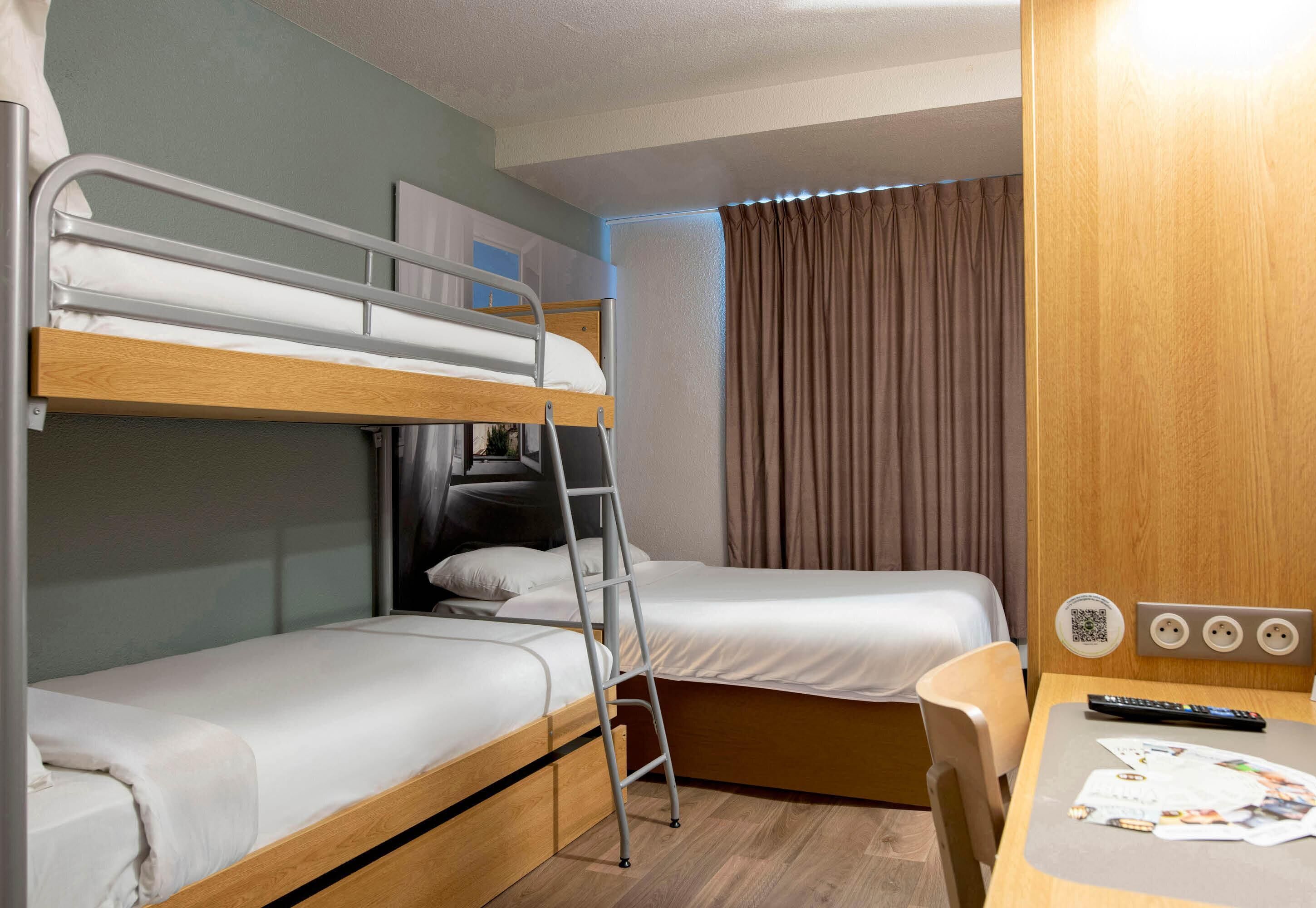 Una habitació d'hotel amb llits superposats, un escriptori amb una cadira, un ordinador portàtil i una finestra amb cortines.