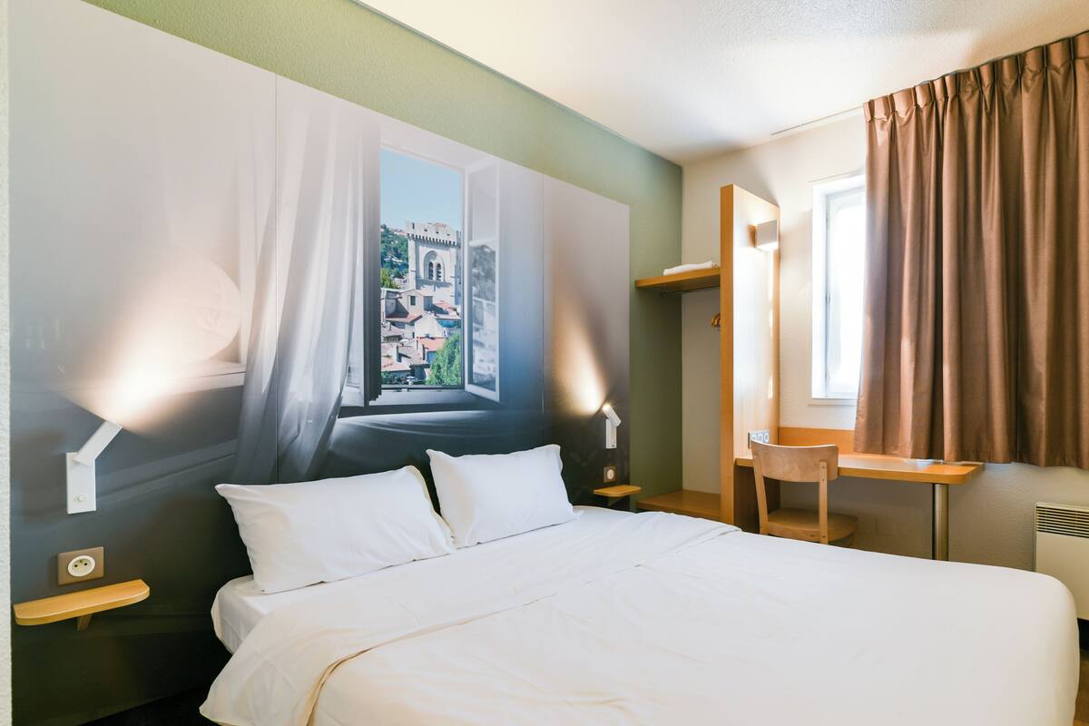 A hotel room with a bed, a desk, a chair, a window with curtains, and a wall mural.
