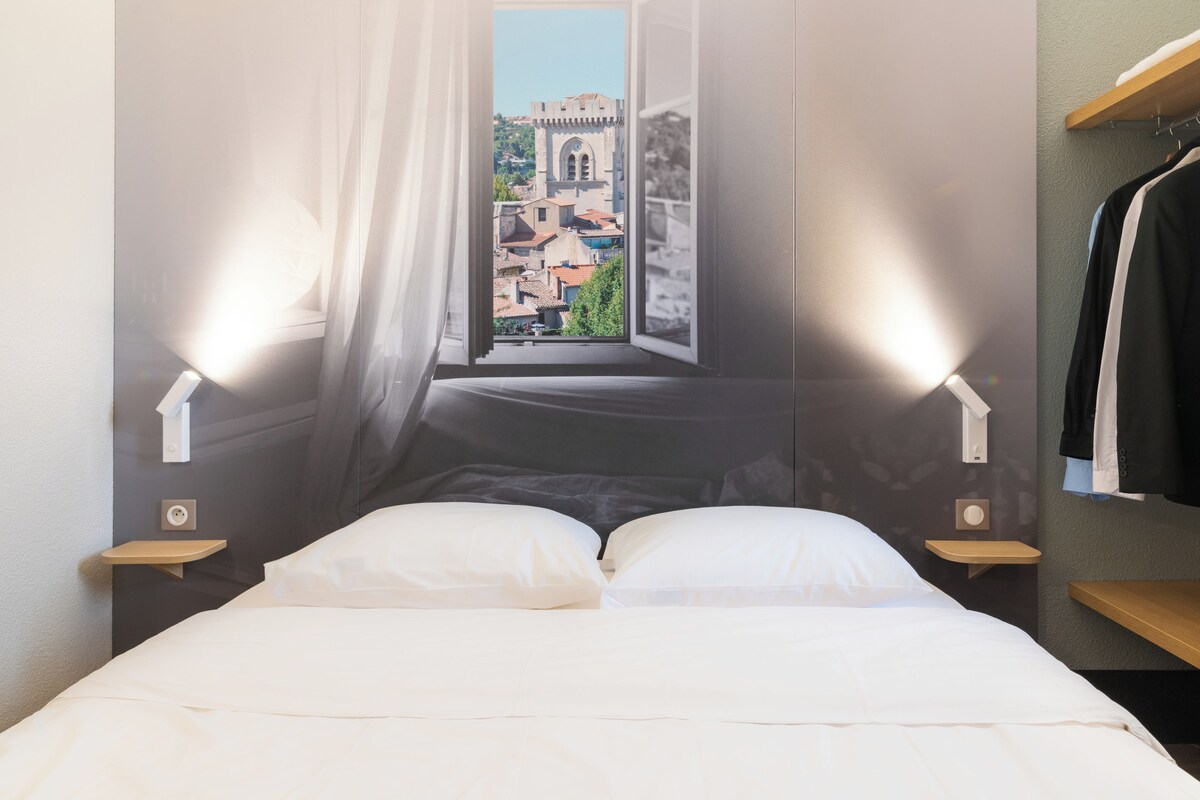 Habitación de hotel con cama, dos mesitas de noche, lámpara colgante y vista a un paisaje urbano a través de una ventana.