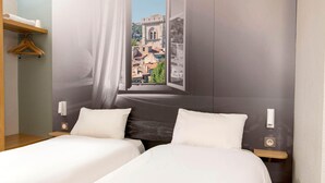 A hotel room with two beds, a window with a view of buildings, a wooden shelf, and a hanging garment rack. - B&B HOTEL Avignon (2) (Le Pontet)