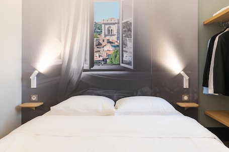 A bed with white linens, a window with a view of buildings, two bedside tables, and a wall-mounted shelf.. B&B HOTEL Avignon (2)