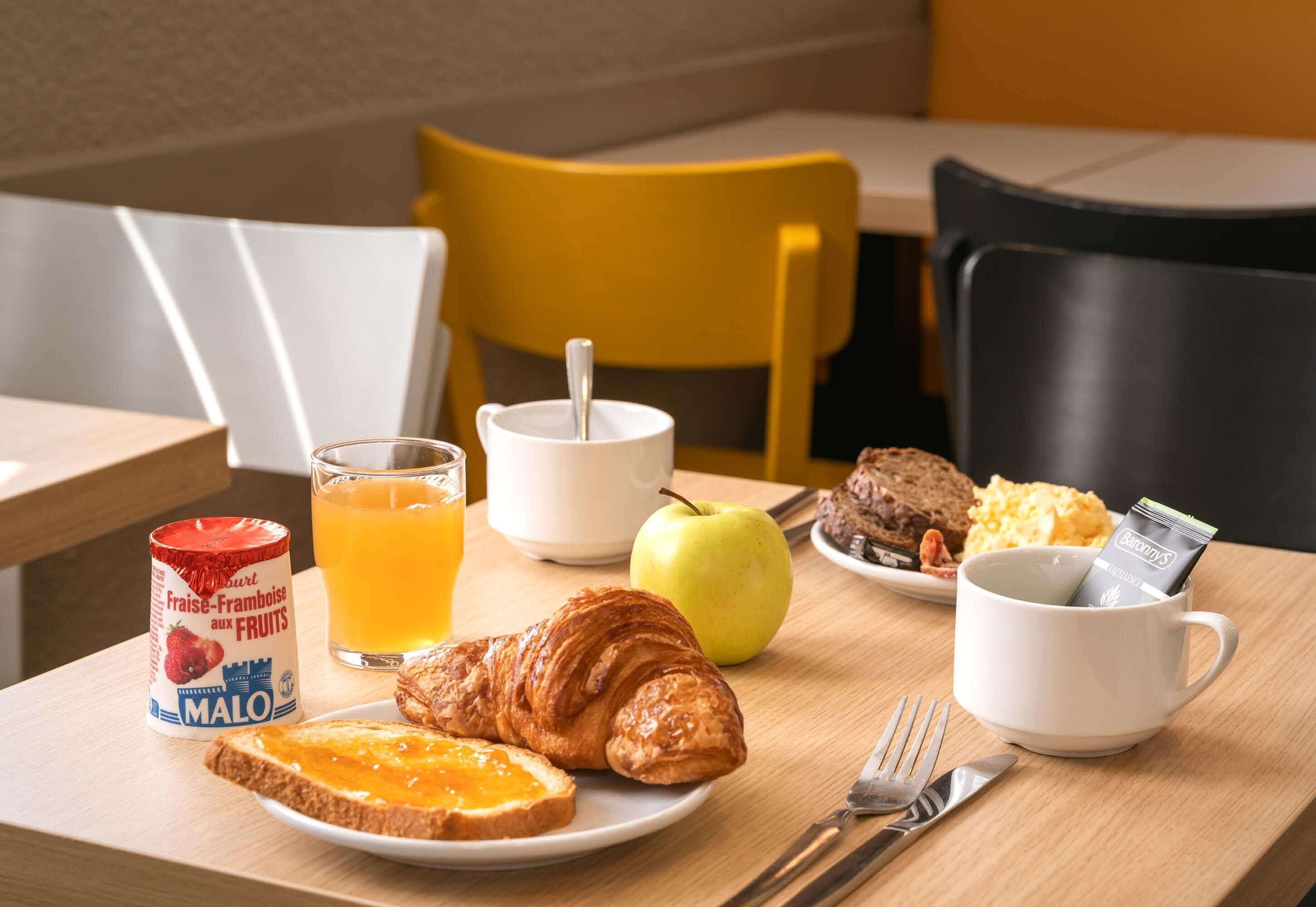 Une table dressée pour le petit-déjeuner avec des croissants, des toasts, un verre de jus d’orange, une tasse blanche, une assiette d’œufs brouillés, une part de gâteau et un pot de confiture de fruits.