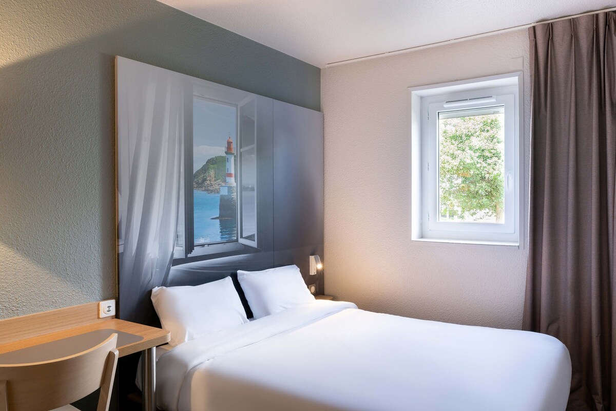 A hotel room with a bed, a desk, a chair, a window with curtains, and a painting on the wall.