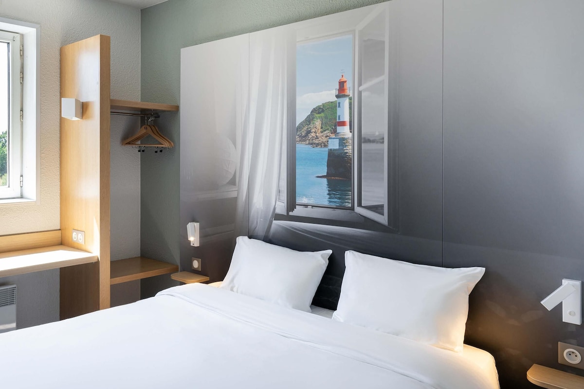 A hotel room with a bed, a window with a view of a lighthouse, a wooden shelf, and a wall-mounted light fixture.
