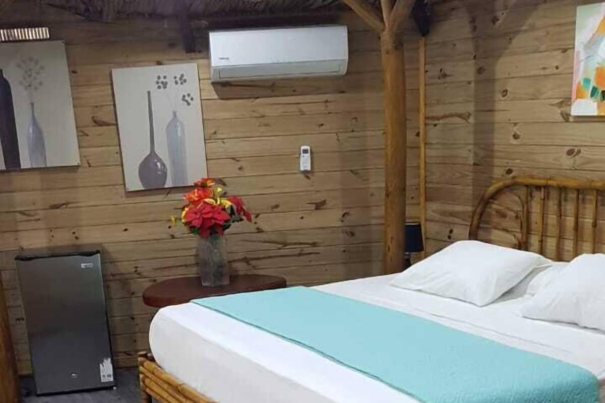 Una habitación con una cama, una mesita con un jarrón de flores, un refrigerador y paredes de madera.