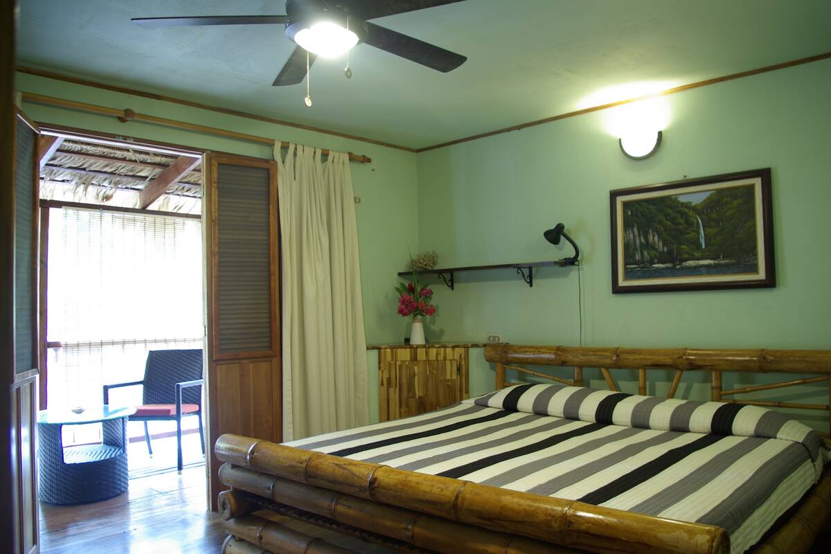 Una habitación con cama, una mesa con sillas, un ventilador de techo y un cuadro en la pared.