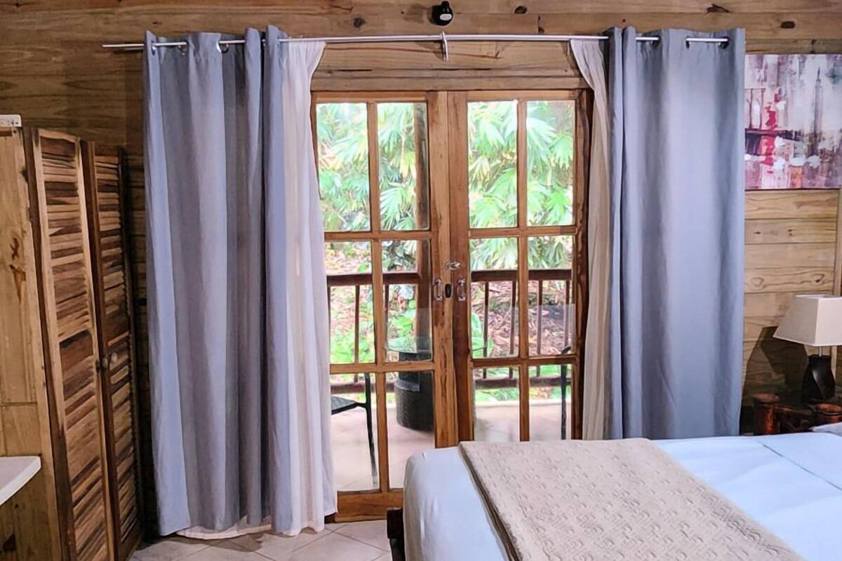 Una habitación de madera con una cama, un ventanal con cortinas y vistas al verdor exterior.