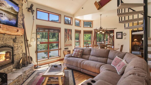 A cozy living room with a stone fireplace, large windows, a comfortable sofa, and a staircase.